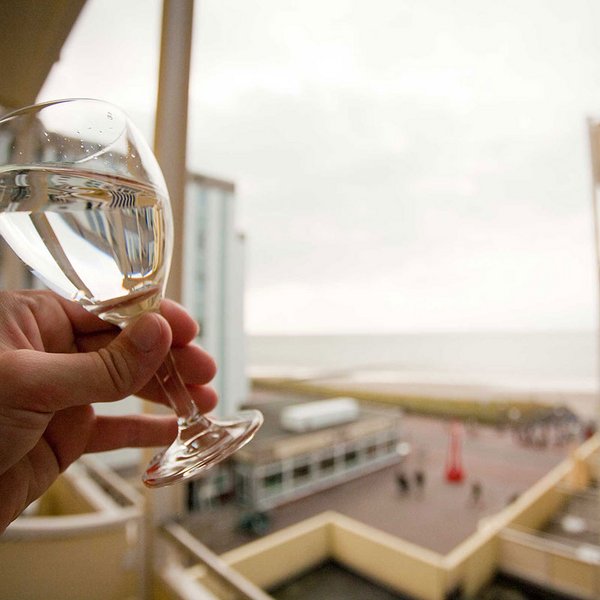 Nahaufnahme von einem Weißweinglas hochgehalten von einer Person, die auf dem Balkon steht. Im Hintergrund die Nordsee und Promenade von Borkum